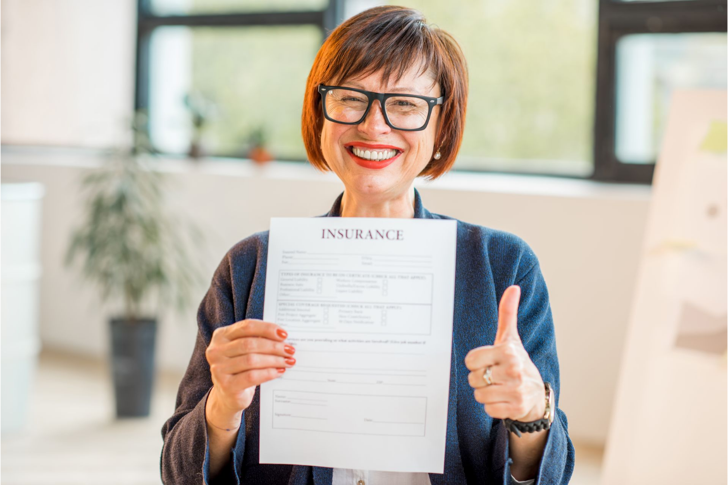 Woman in glasses holds insurance document, smiles, and gives a thumbs up in an office setting.