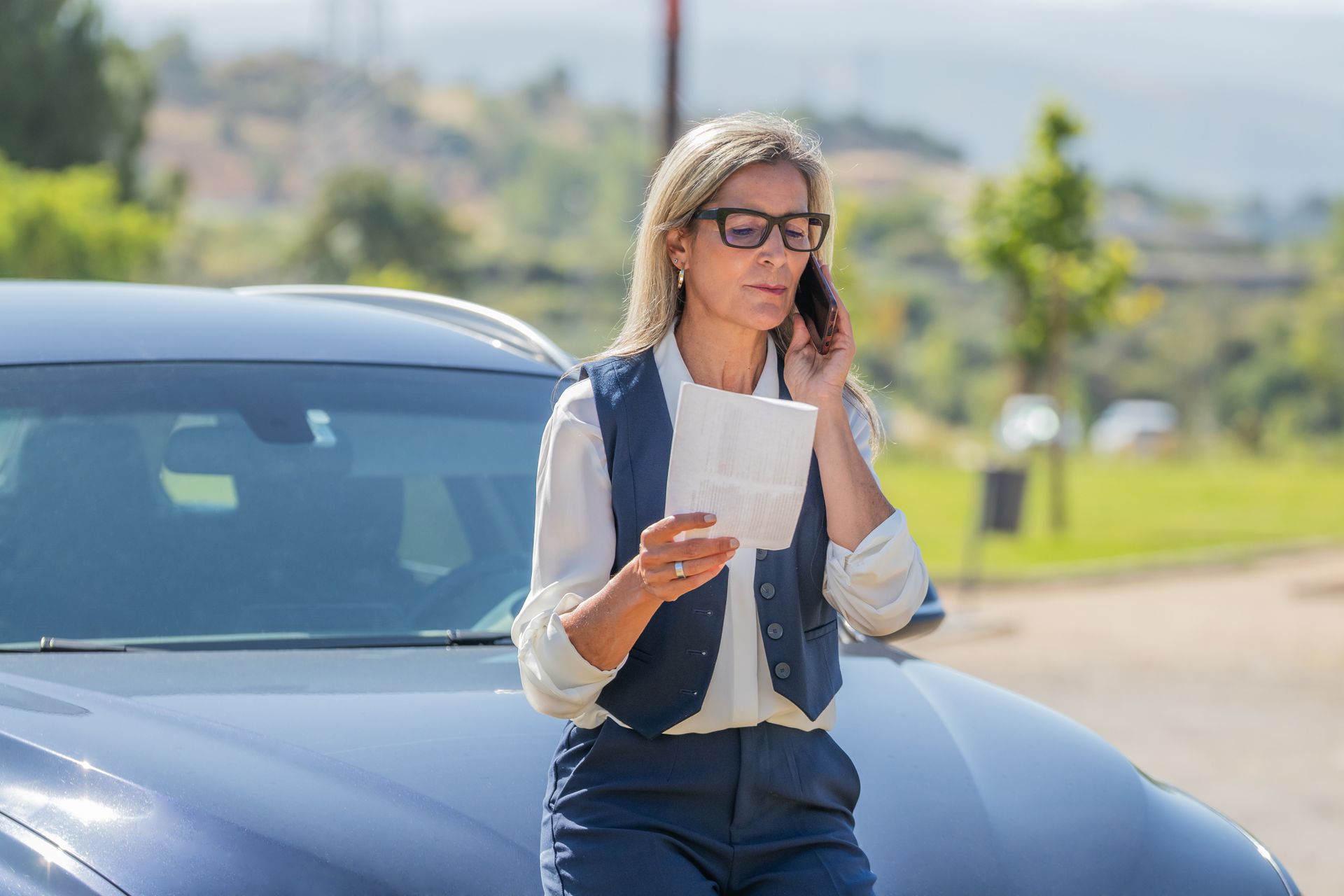 Woman in glasses on phone, reading paper, leaning on blue car in sunlight.