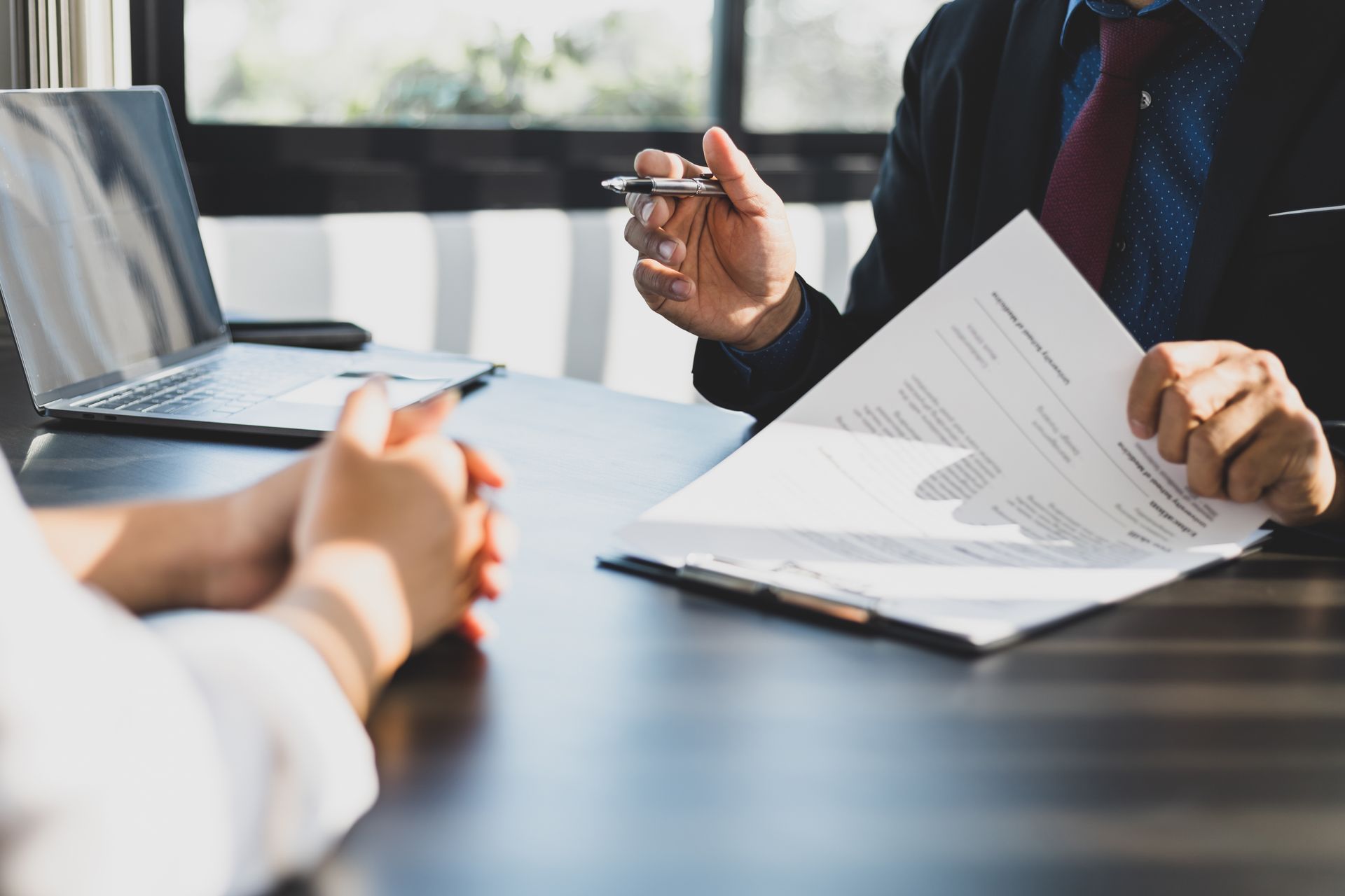 Person in suit points at document during a meeting with another person at a table, a laptop is in the background.