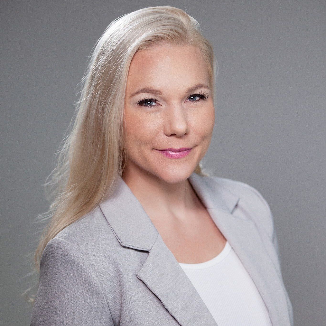 Woman with long blonde hair, smiling, wearing a gray blazer over a white top.
