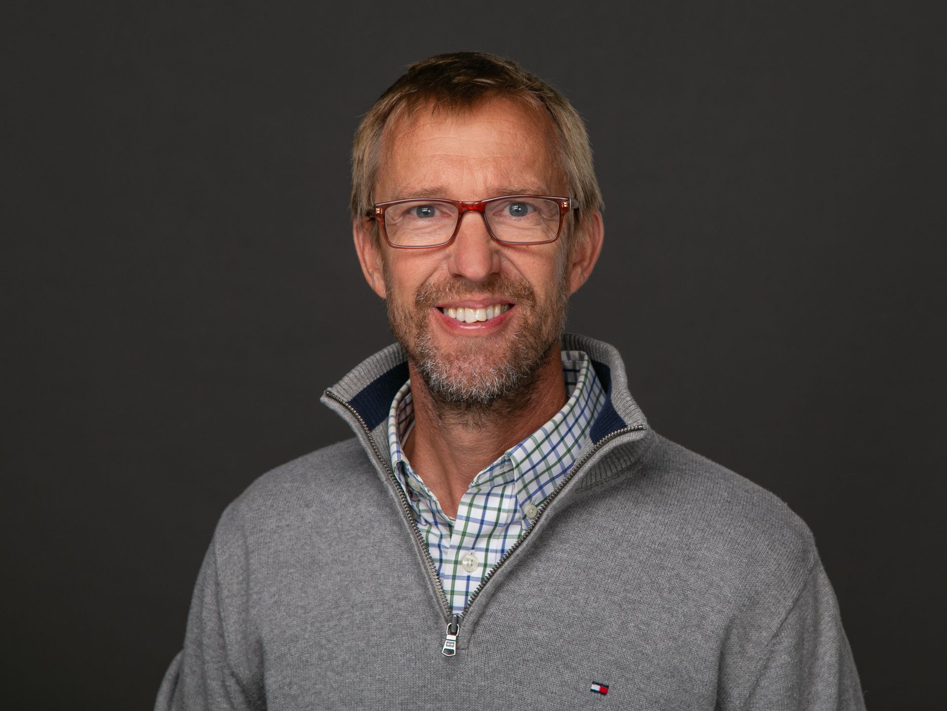 Man wearing glasses and a gray sweater smiles at the camera. Dark gray background.