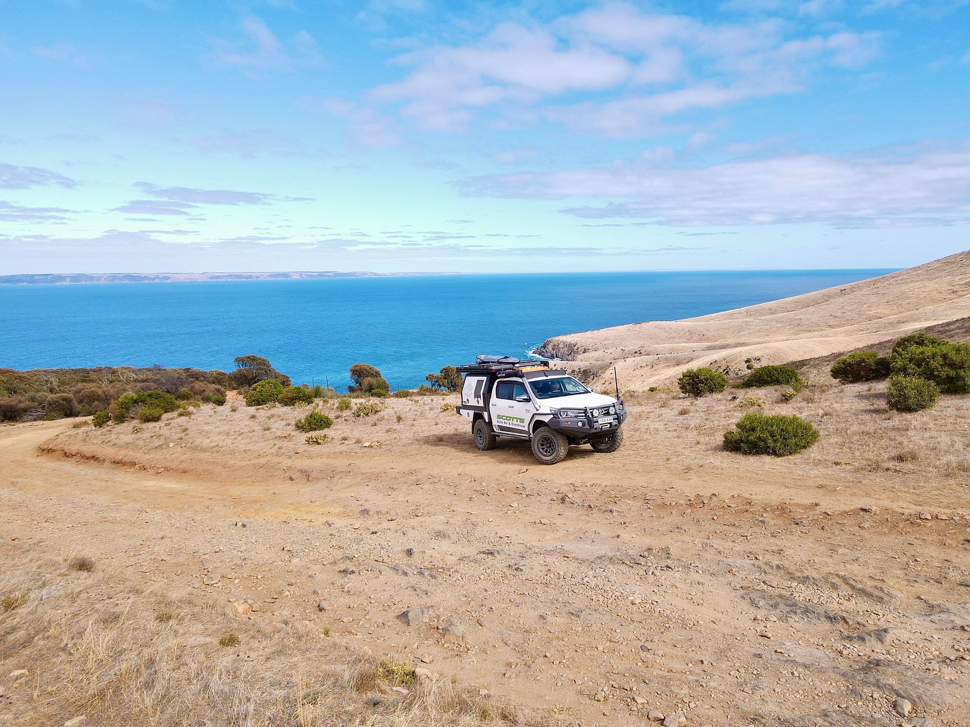 A white ute is parked on a dirt cliff with the ocean behind — Scotts Auto Air & Electrical In Wollongong, NSW