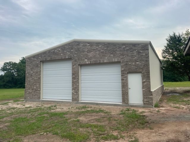 A garage with two white garage doors is sitting in the middle of a grassy field.