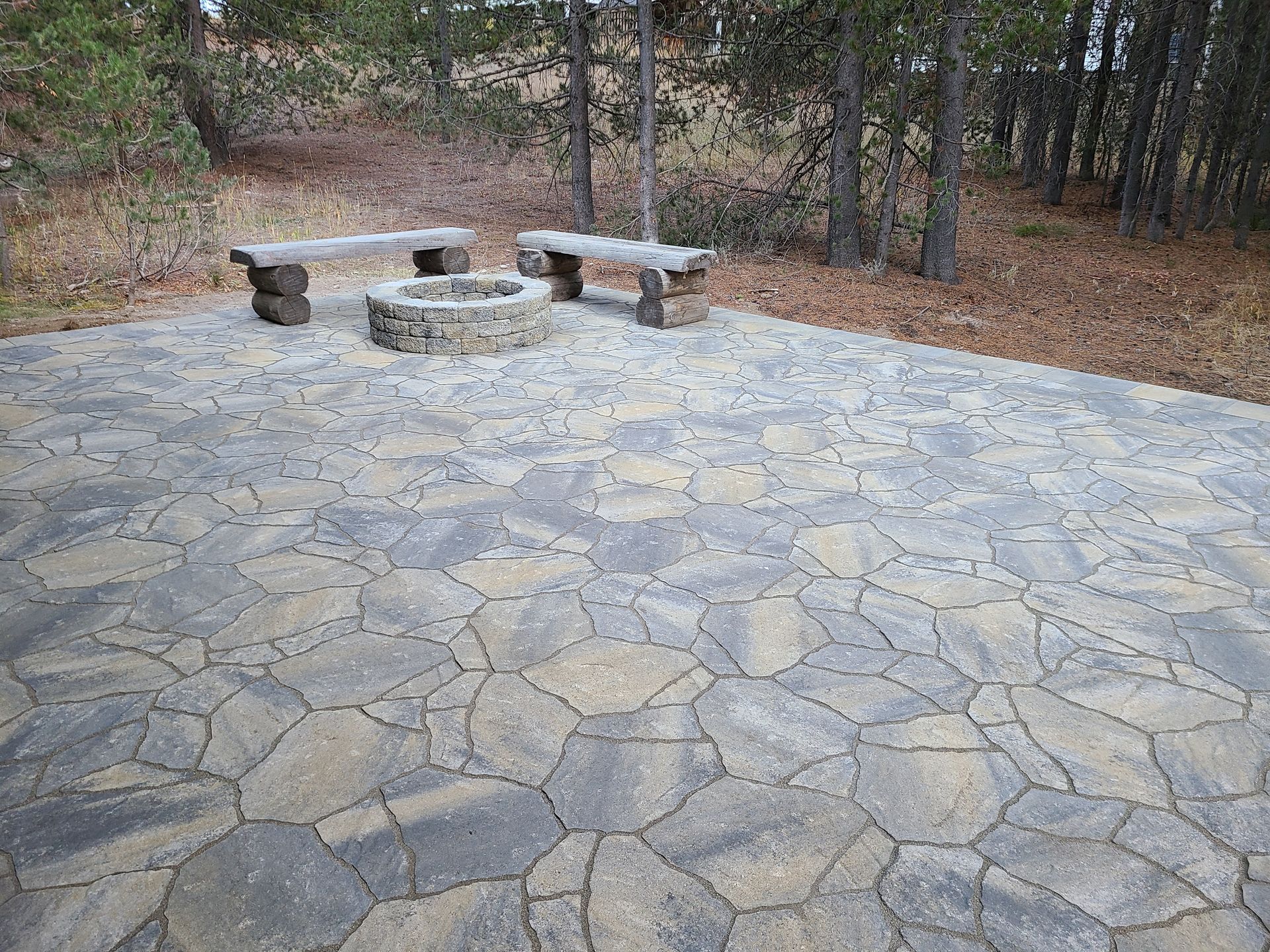 A stone patio with a fire pit and benches in the woods.