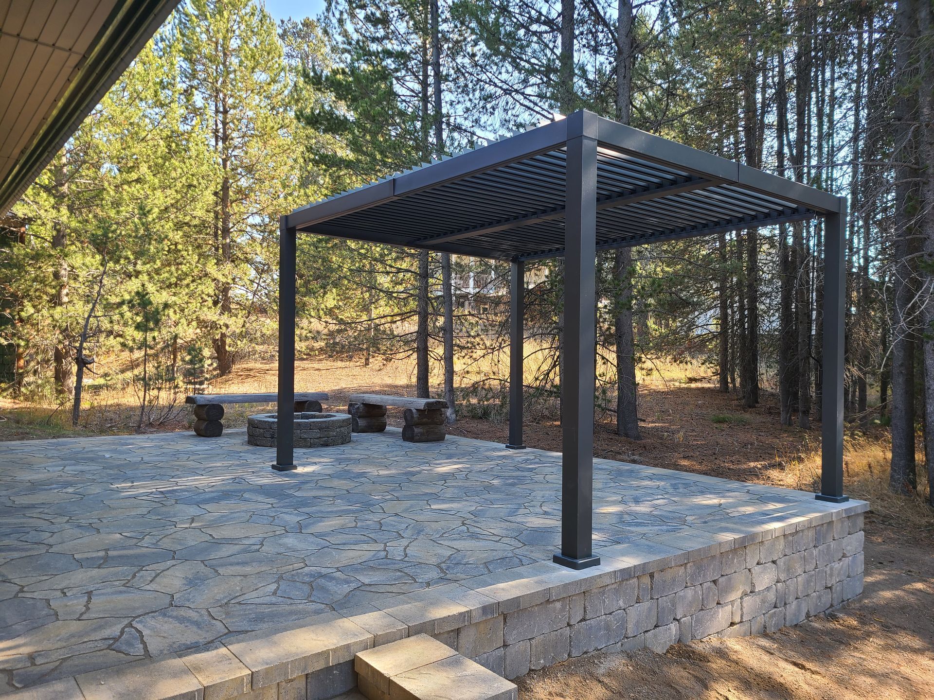 A pergola is sitting on top of a stone patio in the middle of a forest.