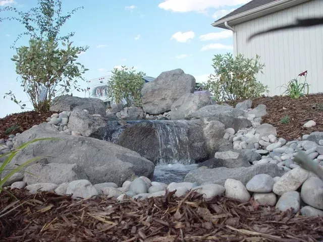 A small waterfall is surrounded by rocks and mulch in front of a house.