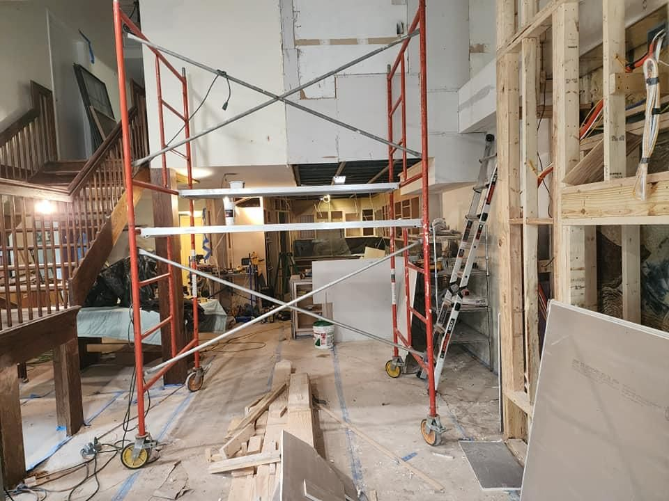 Interior construction site: red scaffolding, exposed wood framing, unfinished walls, debris on the floor.