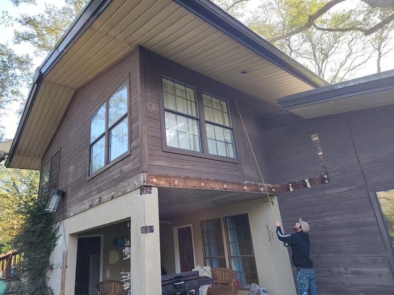 Person attaching string lights to a brown house's porch. Beige and brown siding, sunny outdoor setting.