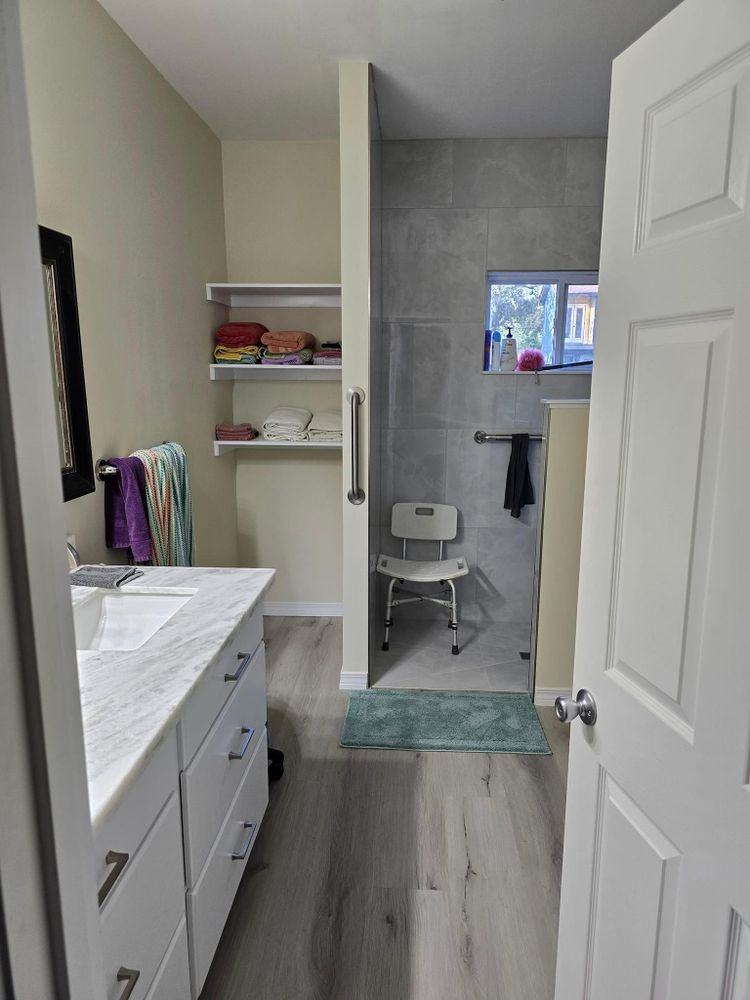 Bathroom with accessible shower, vanity, shelving, and seated shower chair. Light wood floors, white walls.