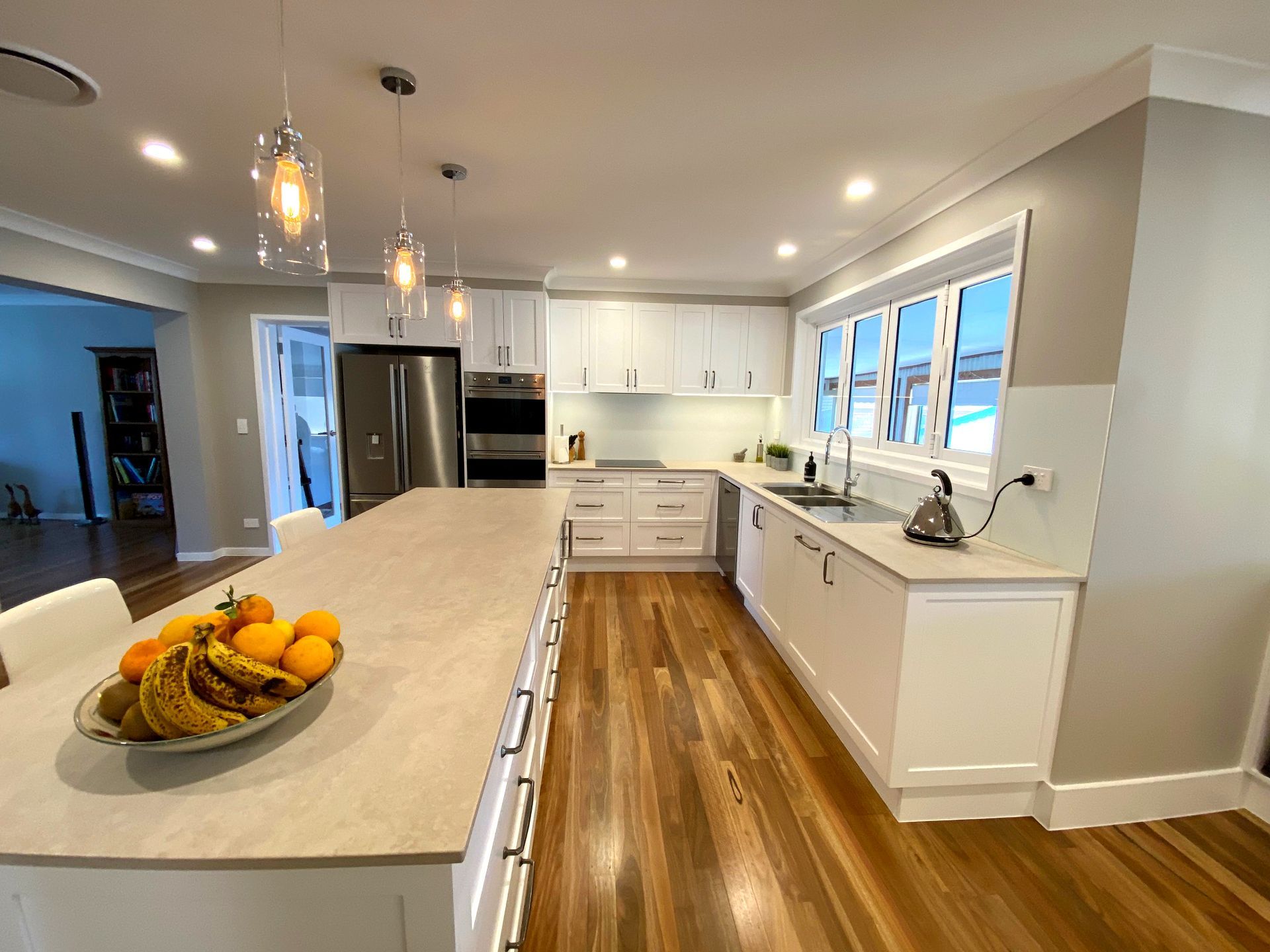 Bright modern kitchen with white cabinets, wood floors, island, pendant lights, and bowl of fruit — BK Building Services Pty Ltd In Forster, NSW