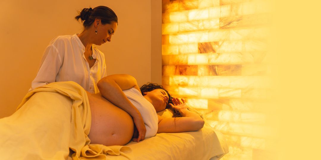 Eine schwangere Frau erhält eine Massage. Ein Therapeut bearbeitet sanft ihren Rücken in einem Spa-Bereich mit Salzziegelwand.