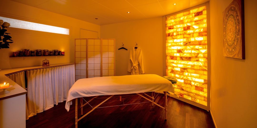 Massageraum mit Salzziegelwand, Massagetisch, sanfter Beleuchtung und Bademantel.