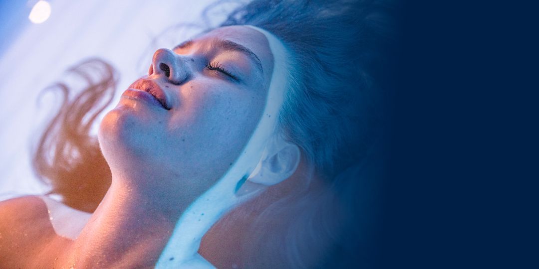 Frau mit geschlossenen Augen, beleuchtet mit blauem und orangefarbenem Licht, liegt in einem Reizentzugstank.