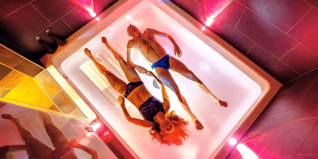 Zwei Menschen schwimmen in einem Wassertank unter rosa Lichtern; die Frau im Bikini, der Mann im Badeanzug.