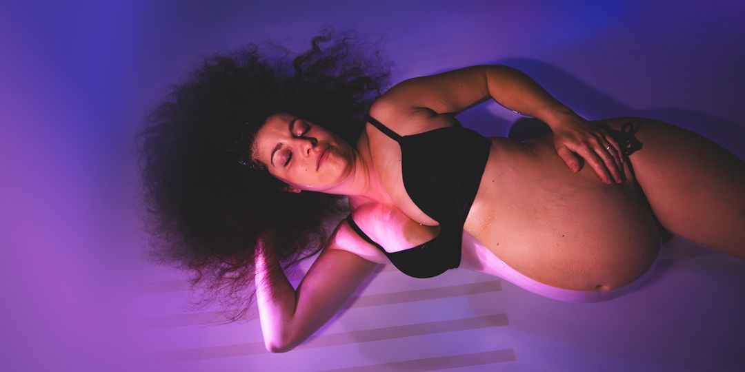 Schwangere Frau mit Afro schwimmt in violettem Wasser, die Augen geschlossen, trägt einen schwarzen BH.