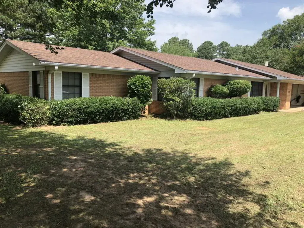 Brick ranch-style house with brown roof and green bushes. Sunny day, with shadows on the grass.