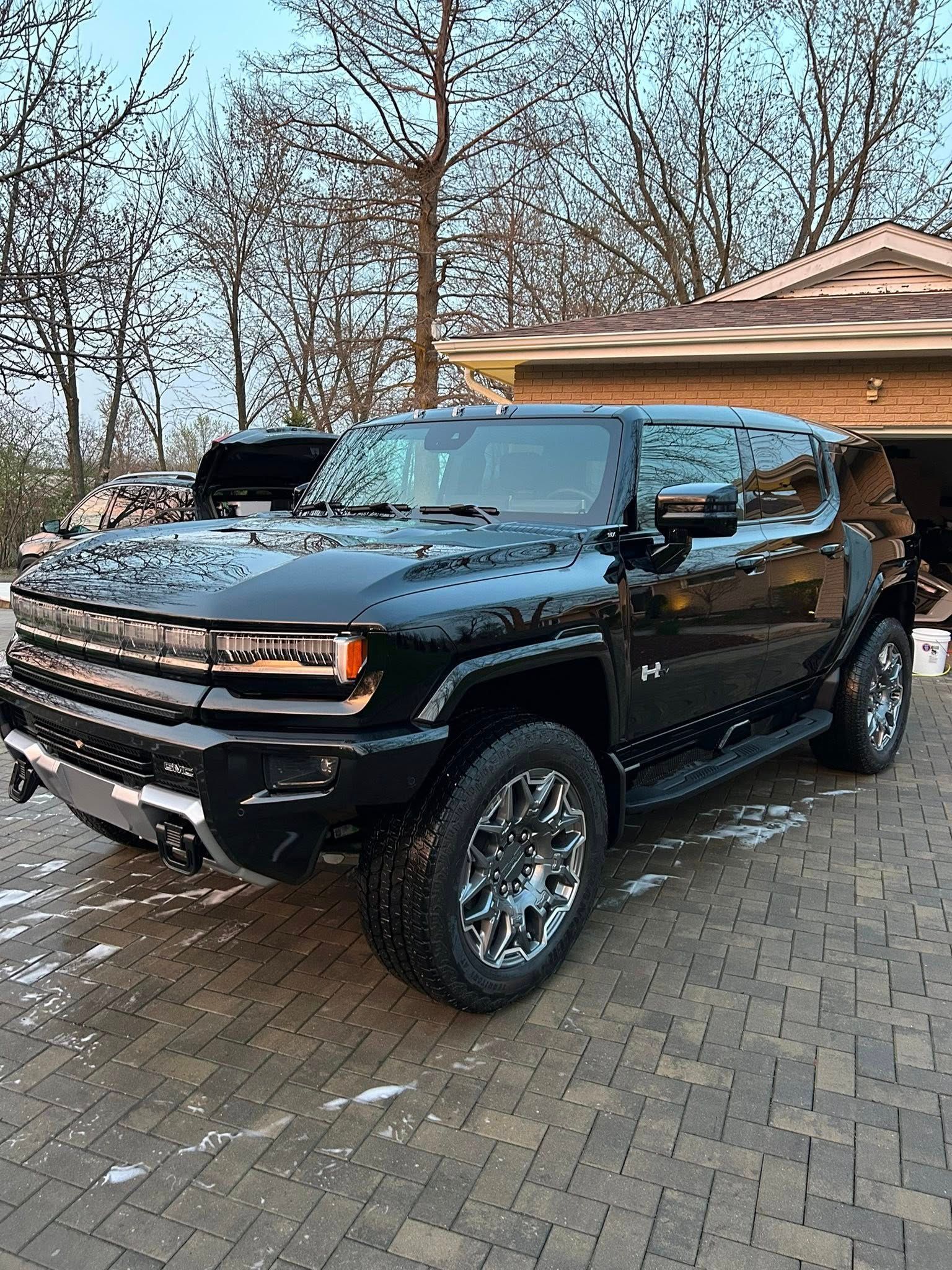 A shiny black GMC Hummer EV pickup truck parked on a paved driveway in front of a house.