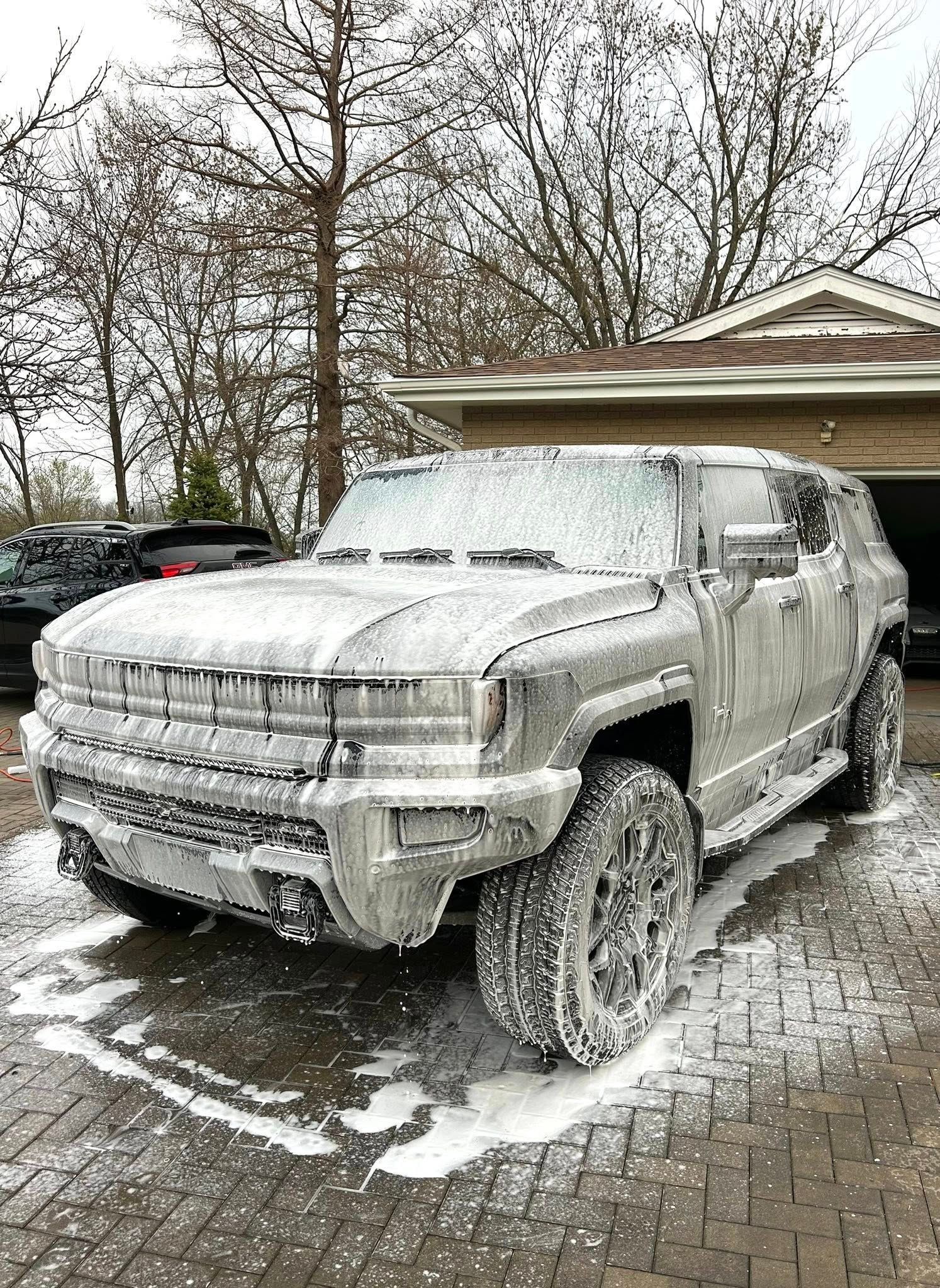 A GMC Hummer EV covered in thick white soap suds, parked on a paved driveway in front of a house.