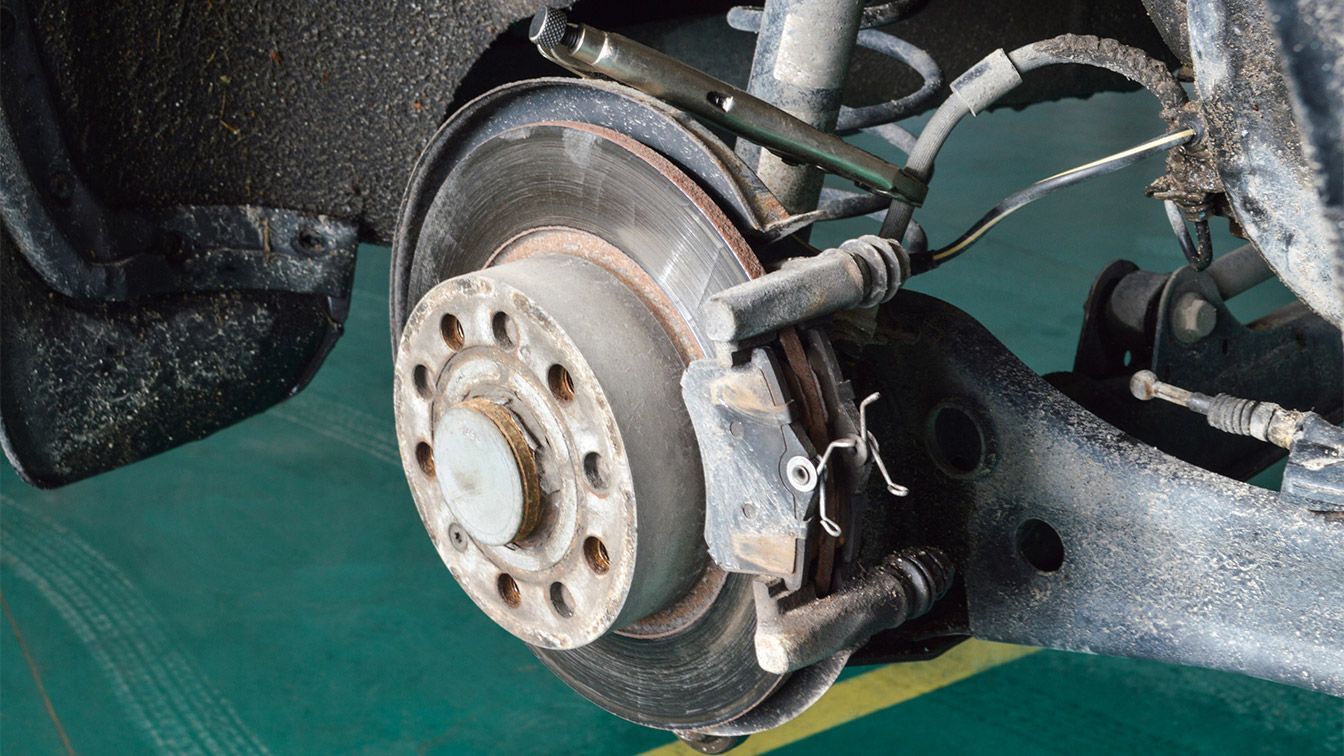 Car's rear disc brake assembly. The rotor, caliper, and suspension components are visible, covered in grime.
