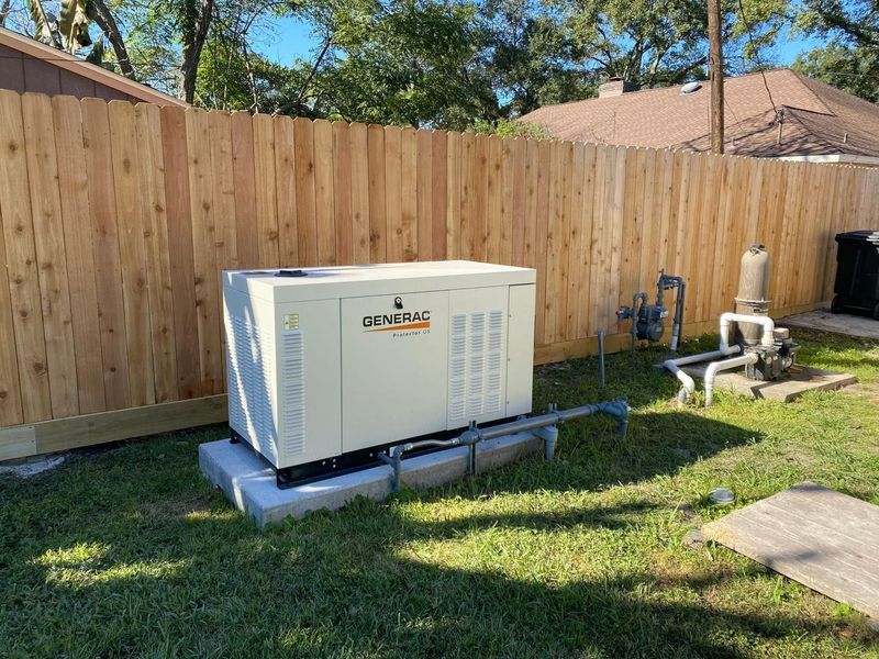 A large, beige generator on a concrete pad in a grassy backyard, beside a wooden fence and pool equipment.