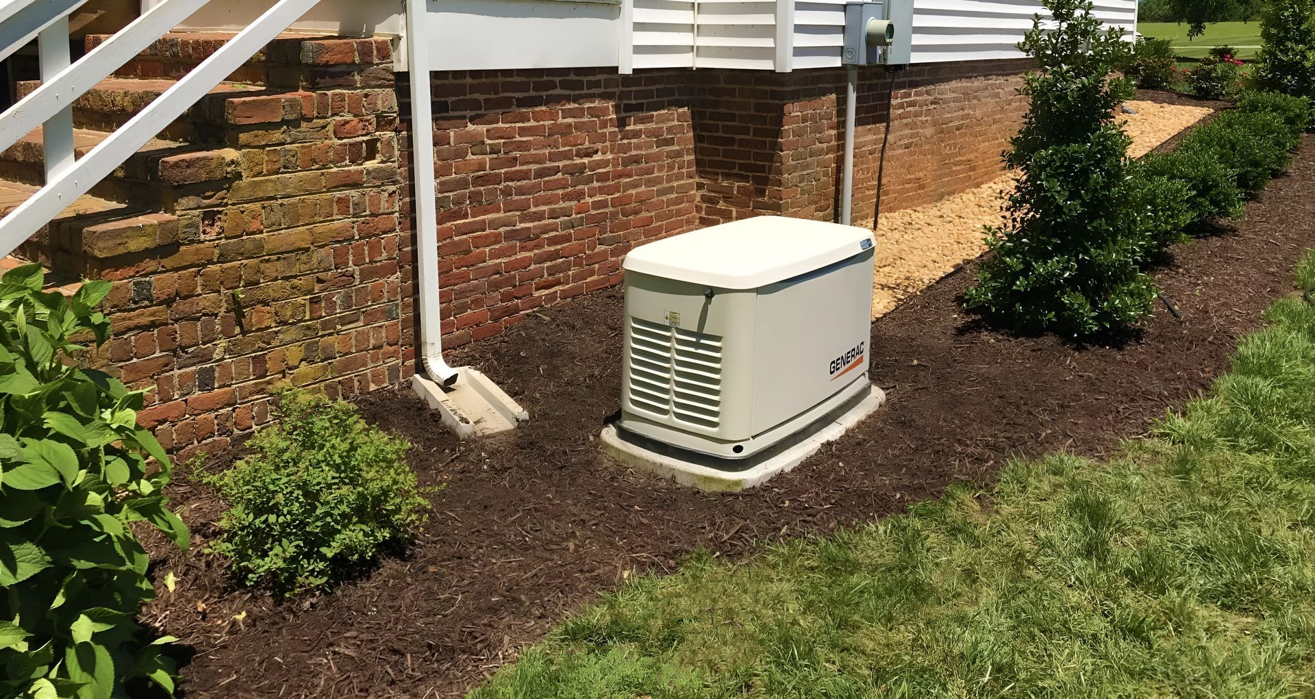 Tan generator near a brick foundation, under white house stairs, on a bed of dark mulch.