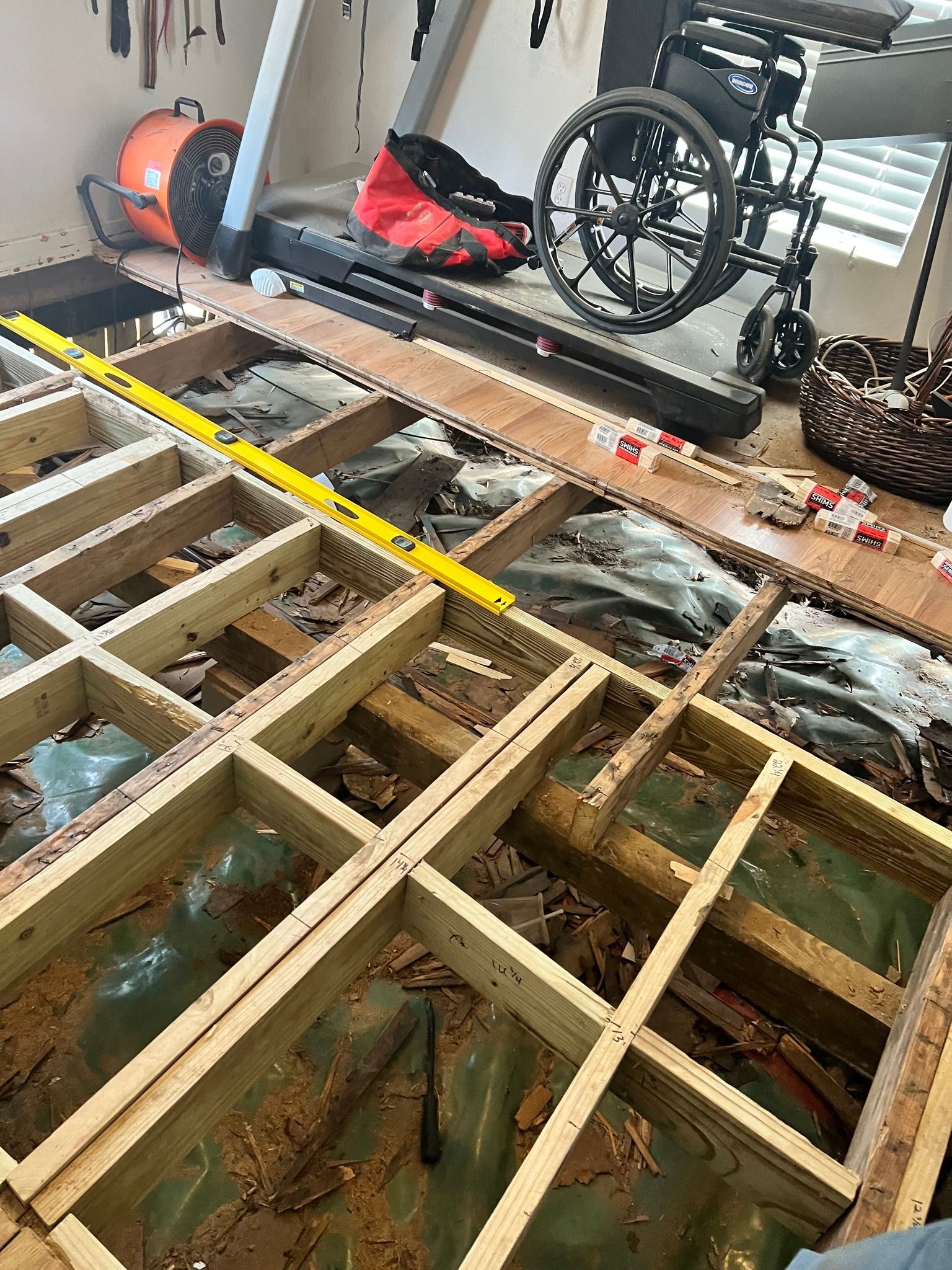 A room with a removed floor, revealing wooden supports. Wheelchair and debris are in the background.