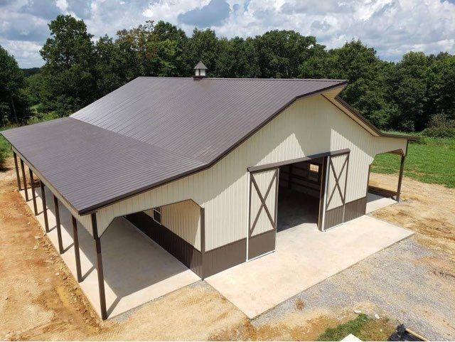 Tan and brown barn with an extended roof, open doors, and a concrete pad.