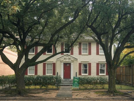 Two-story beige house with red shutters and door, framed by large trees. A pathway leads to the entrance.