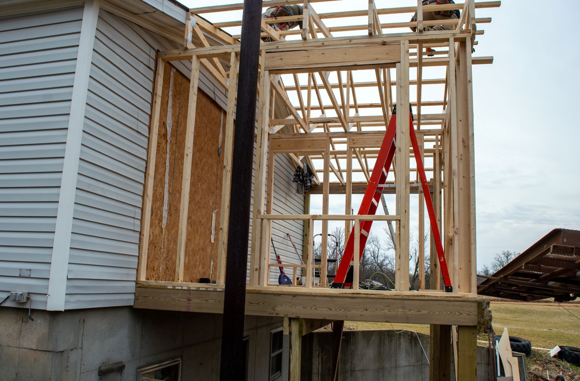 Wooden frame of a house addition under construction, attached to existing siding. A red ladder leans inside.