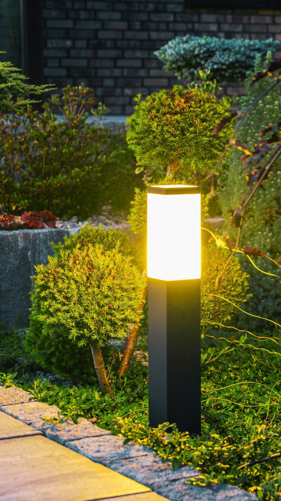 Black outdoor lamp illuminates a garden path with lush green bushes and a brick wall.