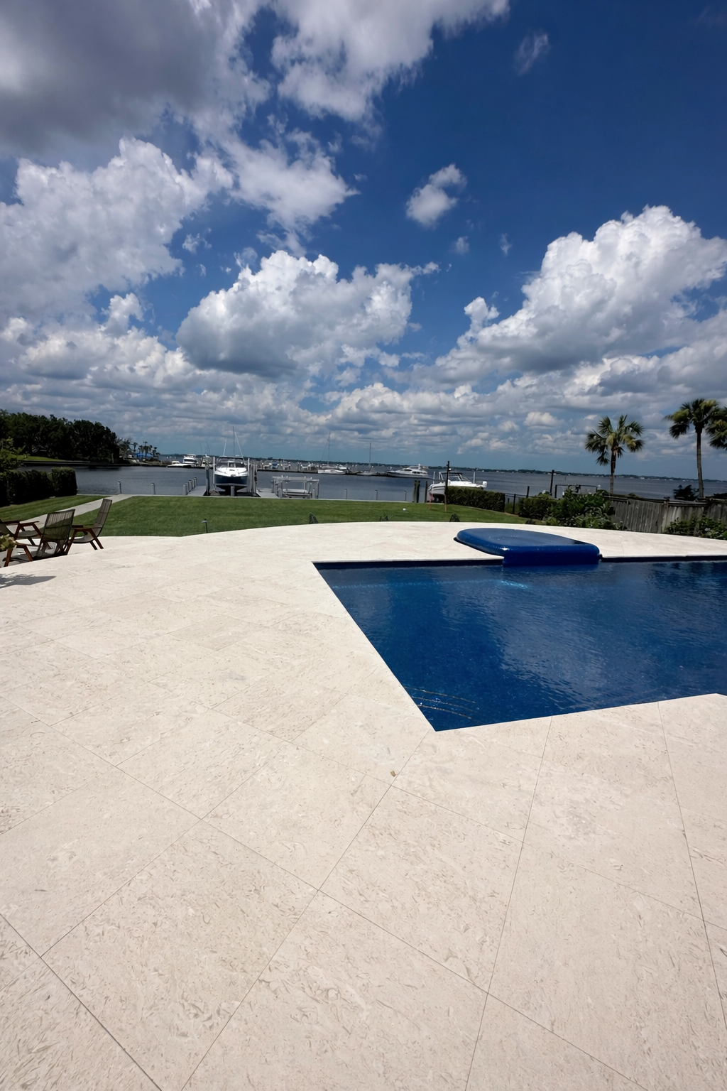 Pool with blue water, white patio, green lawn, harbor, and blue sky with clouds.