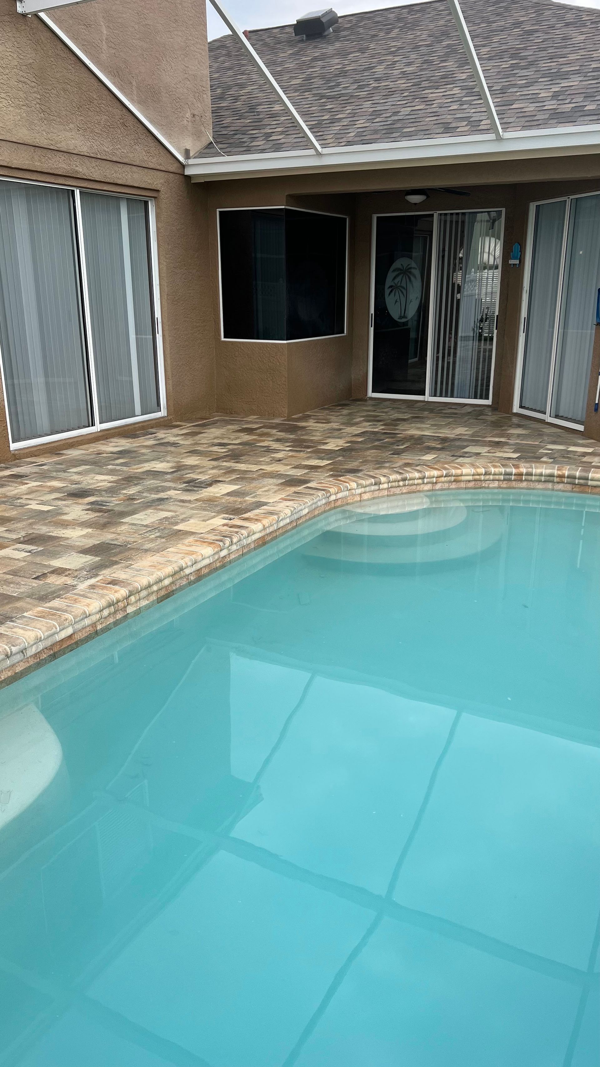 Swimming pool with brick patio, leading to a house with sliding glass doors and a mounted TV.