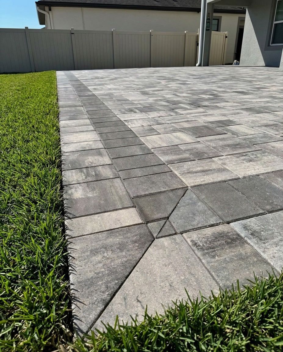 Patio with gray pavers, a dark center border, and green grass alongside a white fence.