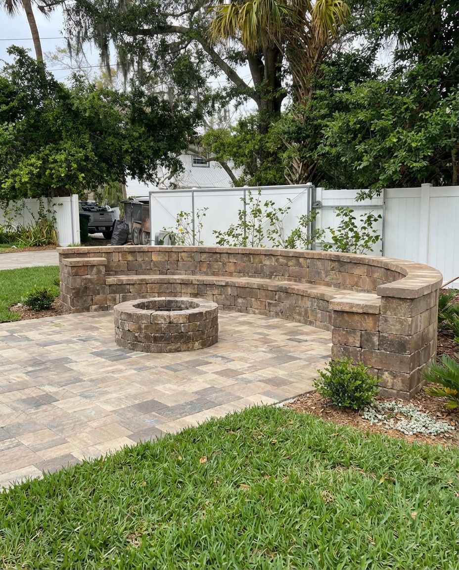 Curved stone seating around a fire pit on a paver patio with a white fence backdrop.