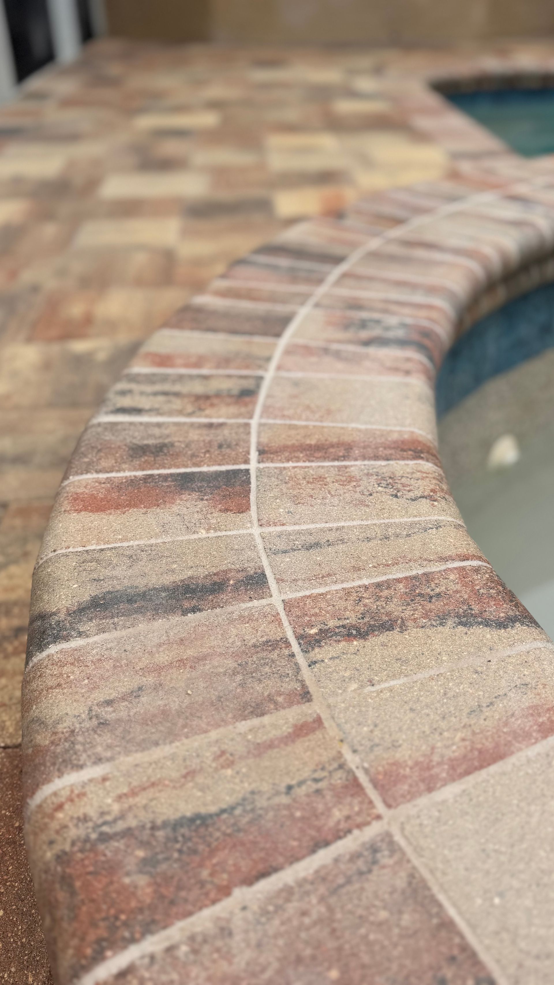 Curved pool edge with textured brick-like tiles, light brown patio in the background.