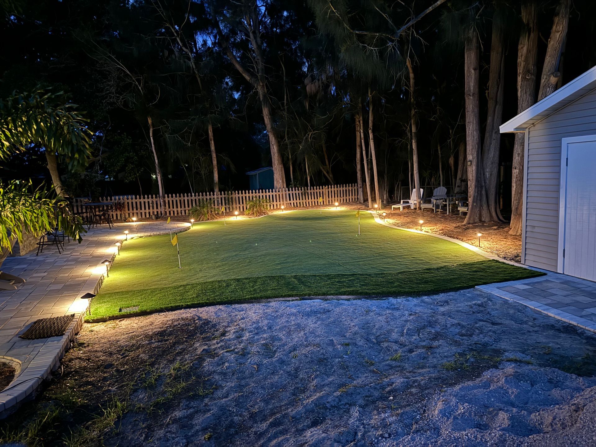 Nighttime outdoor scene with a lit green lawn bordered by landscape lighting, surrounded by trees and a shed.