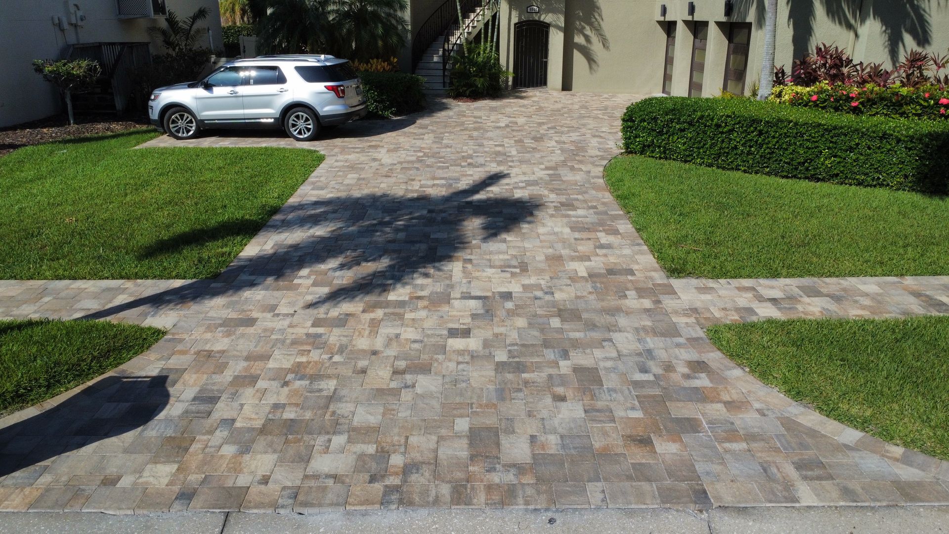 Driveway paved with textured stones, flanked by green grass and a silver SUV parked in front of a house. Palm tree shadow.