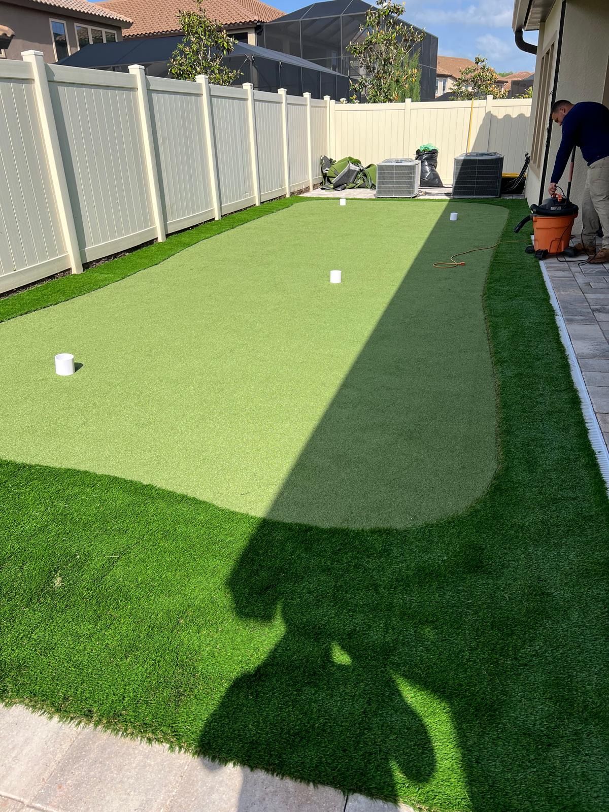 Backyard with artificial turf putting green. Person vacuums the green. White fence surrounds.