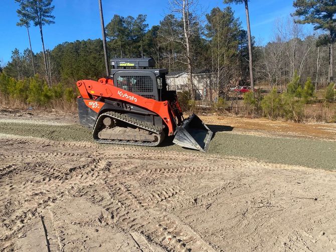Orange Kubota track loader smoothing gravel on a dirt road, trees in the background.