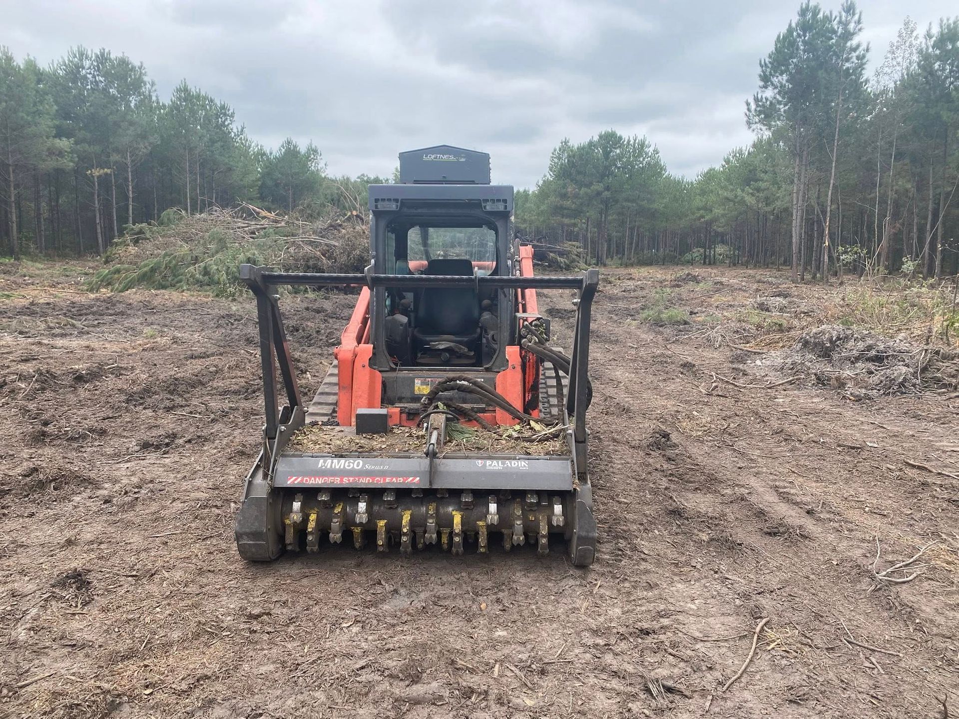 Orange skid steer with mulching head, clearing debris in a forest clearing.