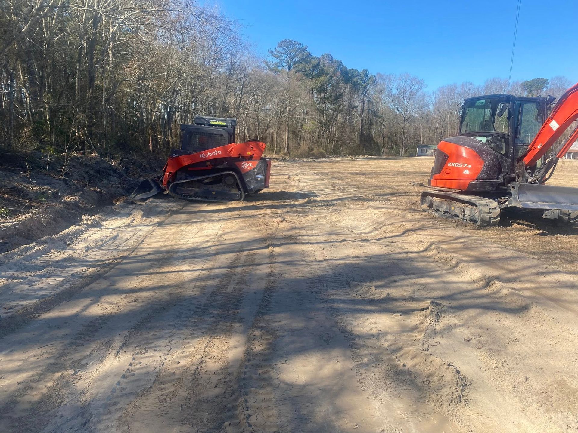 Two orange construction vehicles on a dirt road in a wooded area on a sunny day.
