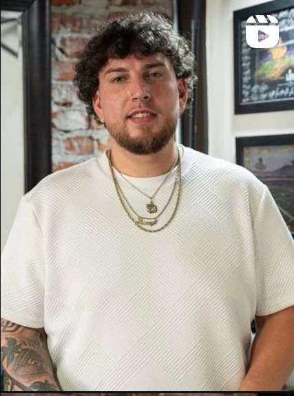 A person with curly hair and a beard wears a white textured t-shirt and layered gold necklaces against a brick wall.