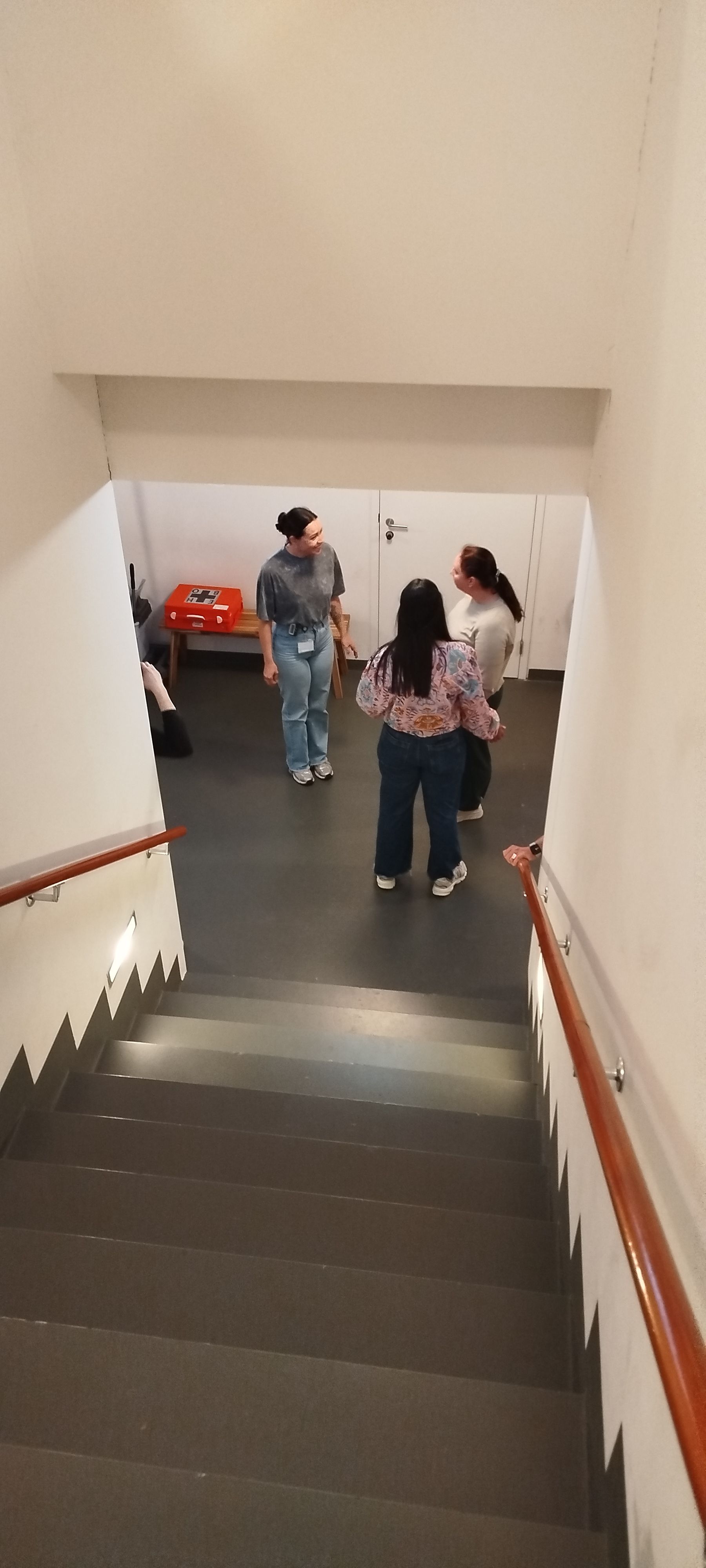Drie mensen praten in een trappenhuis. De persoon vooraan kijkt weg, twee anderen zitten tegenover elkaar.