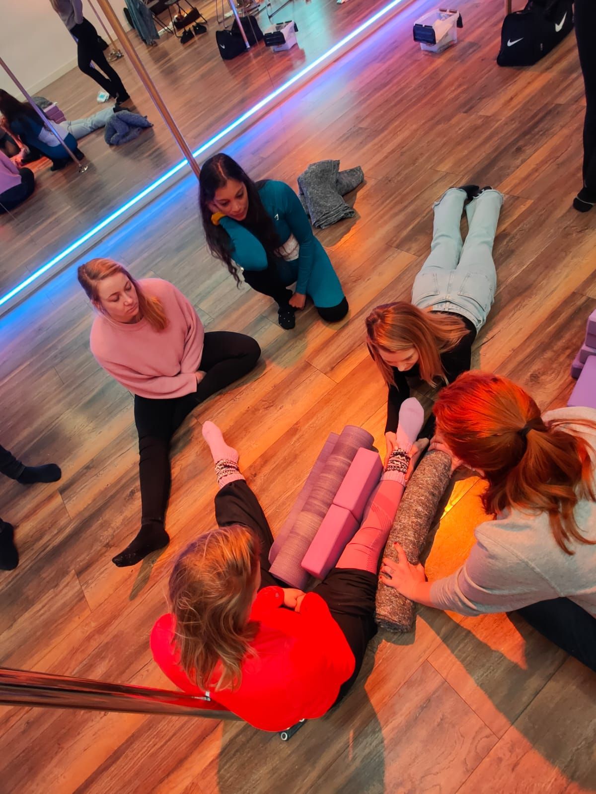 Mensen in een dansstudio met yoga-accessoires; één persoon in het rood ligt op de grond, anderen staan ​​eromheen.