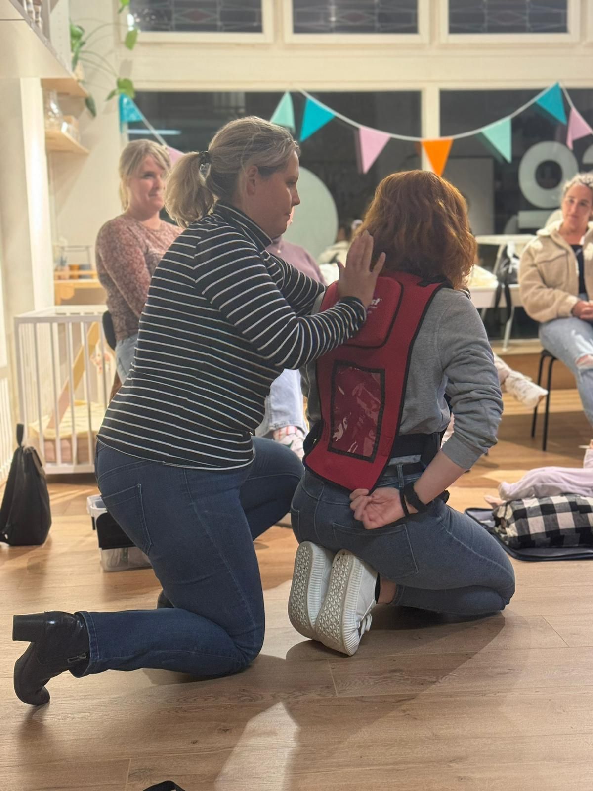 Vrouw demonstreert verstikkingshulp bij een persoon die een trainingsvest draagt. Binnen, in het bijzijn van toeschouwers.