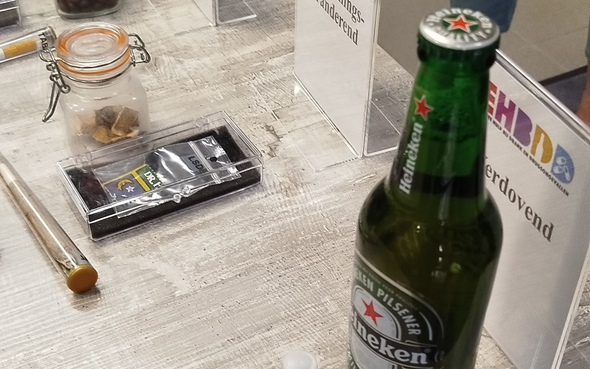 Een groene Heineken-bierfles naast andere spullen op een tafel, waaronder een pot en verpakkingen.