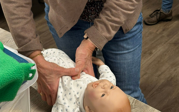 Persoon demonstreert reanimatie bij een baby op een oefenpop. Handen op de borst, binnenomgeving.