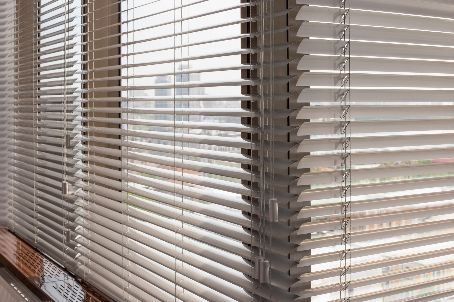 White, slatted blinds in a corner window, filtering sunlight and obscuring a city view.