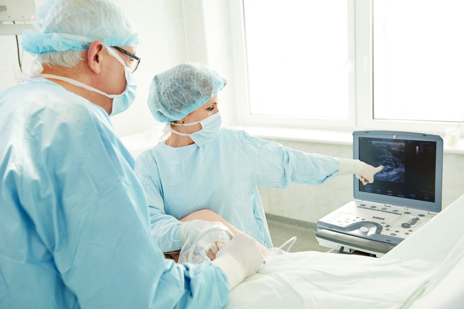 Two medical professionals in surgical scrubs point to an ultrasound screen while examining a patient in a bright room.