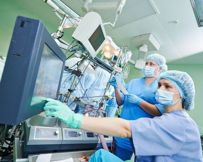 Two medical professionals in scrubs and masks monitor data on a screen in a clinical setting.